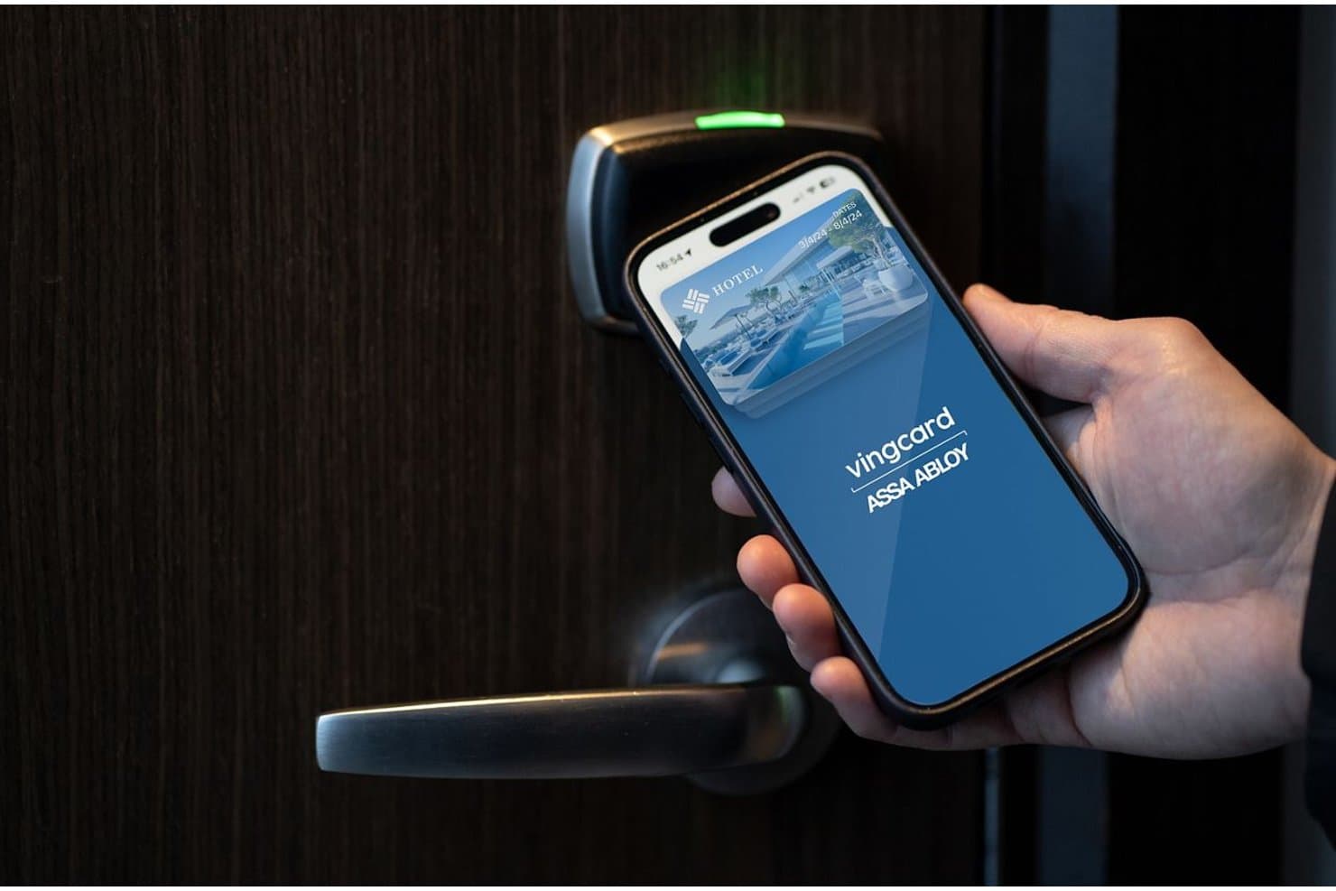Hand holding smartphone with Vingcard app near electronic hotel door lock.